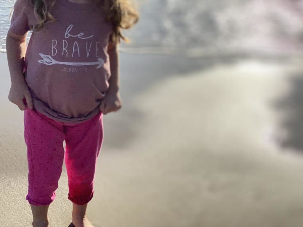 A child at the beach wearing a “be brave — Joshua 1:9” shirt